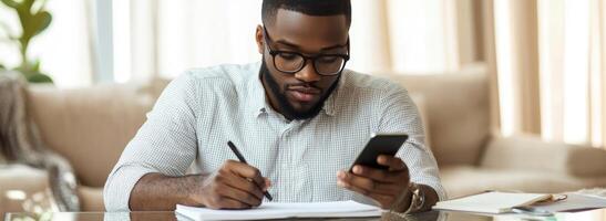A man with glasses and a beard is writing on a notepad while looking at his phone photo
