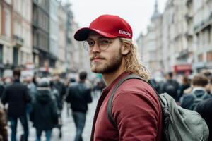 Young man with glasses and long hair strolls through a busy European street, showcasing a vibrant urban atmosphere during a lively afternoon photo