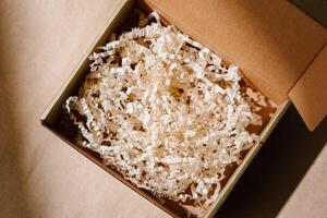 Open cardboard box with shredded paper packaging resting on a light brown surface illuminated by soft natural light photo