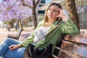 Woman looking at the camera while relaxing on a bench in a sunny park with blooming purple trees photo