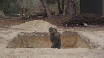 A man wearing a hat and jacket is digging a deep hole in the sandy ground. Clumps of soil are being thrown aside as he works diligently on this excavation project. video