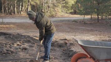 A man in a green jacket and cap uses a shovel to shift soil into a wheelbarrow at a construction site video