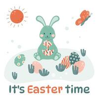 Easter bunny is sitting on the grass with colorful eggs and holding an Easter egg in his paws. vector