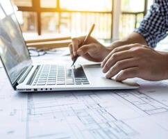 Professional engages in design work while using a laptop on construction blueprints in a bright modern workspace photo