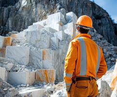 Worker observes marble quarry operations under bright blue sky in early afternoon light near mountainous terrain photo