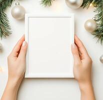 blank white frame mockup held by hands, surrounded by festive Christmas decorations like ornaments and pine branches, creating cheerful holiday atmosphere photo