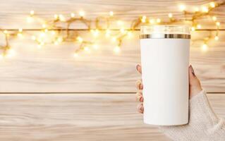 person holding stylish white tumbler against backdrop of warm fairy lights, creating cozy atmosphere. colorful nails add playful touch to scene photo