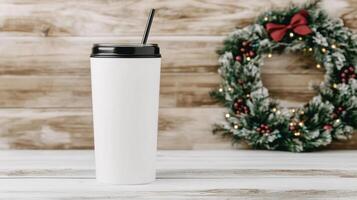 blank white tumbler mockup stands against festive wreath backdrop, perfect for holiday promotions. Ideal for showcasing designs or branding photo