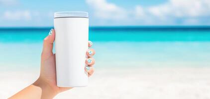 hand holding white tumbler against beautiful beach backdrop, featuring turquoise water and clear sky. scene evokes sense of relaxation and enjoyment photo