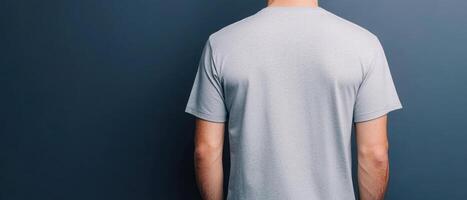 man wearing grey t shirt stands against dark blue wall, showcasing back view of shirt. simple design highlights fabric texture and fit, creating casual and modern look photo