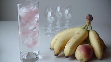 A tall glass filled with ice and a hint of pink sits beside ripe bananas and a peach photo