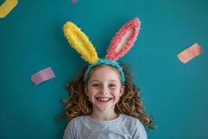 A joyful girl wearing colorful bunny ears smiles on a bright turquoise background with playful decorations around her photo