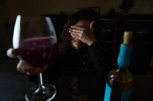 Woman with wine at table, covering face in dark, contemplative mood photo