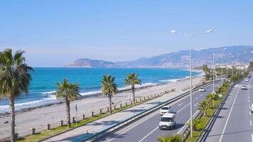 Vehicles passing on a highway from a pedestrian bridge besides the beach and sea showing the blue sky and sea waves crashing on the sandy beach in Alanya, Turkey during a sunny day video