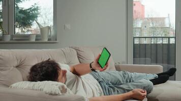 Man in casual clothes lies on beige couch, holding smartphone with green screen, relaxing in modern living room. Male resting on sofa and watching online on mobile phone with chroma key display video