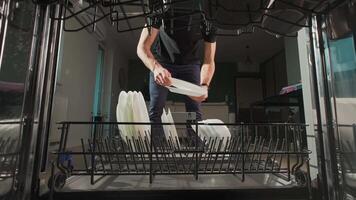 Man unloading clean white plates from the bottom rack of dishwasher in kitchen. Person performing everyday household chores, using modern home appliance video