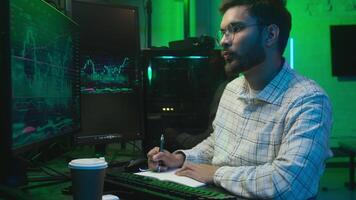 Indian Arabian man writing notes notebook computer pc monitor freelancer crypto trading chart screen male programmer guy IT broker analyst write bitcoin online currency working results neon e-commerce video