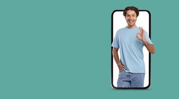 A young individual wearing a light blue shirt stands confidently while displaying an okay gesture. The large smartphone screen emphasizes their friendly demeanor against a clean background. photo
