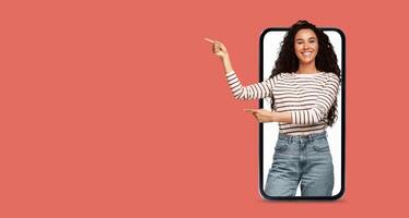 A young woman with long, curly hair is smiling and pointing at a large smartphone screen. The background is a solid, vibrant color, creating an engaging communication atmosphere. photo