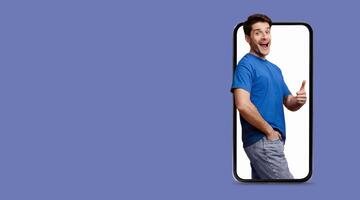 A cheerful young man is interacting with a large smartphone screen, giving a thumbs up while dressed casually. He's surrounded by a solid purple background, creating an upbeat and modern atmosphere. photo