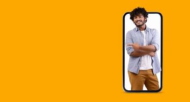 A cheerful young man smiles and poses with crossed arms while appearing to interact with a large smartphone screen in a vibrant yellow background. The scene focuses on modern communication. photo