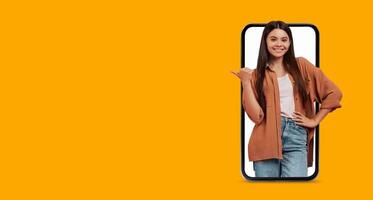 A woman stands confidently, smiling and pointing to her smartphone with a large screen. The bright background enhances the technology and communication aspects of the scene. photo