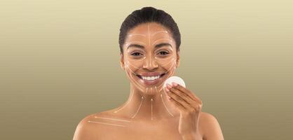 African American woman is engaged in her beauty routine, smiling while applying skin care products. She is in a calm, isolated setting with a neutral background, emphasizing self-care and pampering. photo