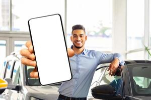 Wealthy young middle eastern man posing by brand new black sports car, showing smartphone with empty screen and smiling at camera, mockup, renting auto on Internet, copy space. Car dealership online photo