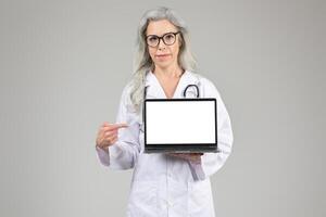 A middle aged woman wearing a white lab coat and glasses is pointing at a laptop with a blank screen. She is displaying a professional demeanor, emphasizing the importance of healthcare technology. photo