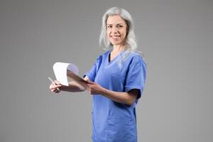 A middle aged woman dressed in blue scrubs is smiling as she holds a patient chart while standing against a neutral background. Her confident demeanor reflects her dedication to healthcare. photo