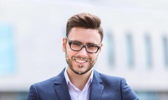 A man wearing glasses and a suit smiles confidently while standing outside. The background is blurred, highlighting his face and the facial recognition grid overlaid on it. photo
