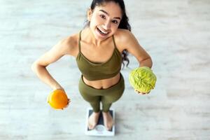 Above view of happy Indian lady holding citrus fruit and cabbage, standing on scales, choosing healthy diet, full length. Young Hindu woman eating balanced raw vegan meals. Wellbeing and weight loss photo