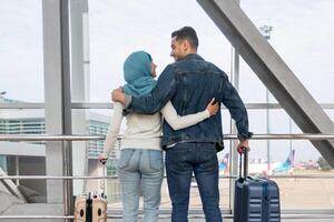 Romantic Travel. Loving Muslim Couple Standing With Suitcases Near Window In Airport, Happy Islamic Spouses Embracing And Smiling To Each Other, Ready To Honeymoon Trip, Rear View With Copy Space photo