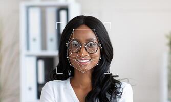 A woman with glasses smiles confidently while facial recognition technology analyzes her features. The modern office backdrop emphasizes innovation and professional atmosphere. photo