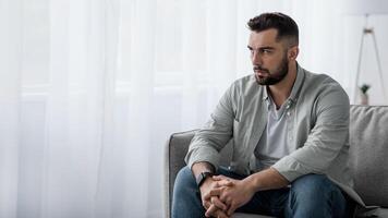 Thoughtful serious bearded millennial male sit on sofa alone at home, lost in thoughts, thinking about problem solving, feeling lonely, make important decision, have psychological problem, side view photo