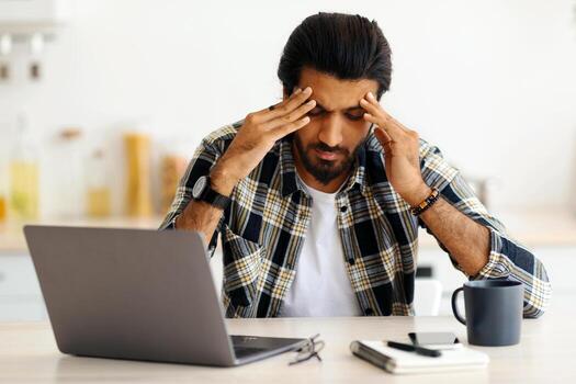 agotado indio hombre sentado en frente de ordenador portátil a hogar, conmovedor su cabeza, sufrimiento desde agotamiento o financiero crisis, joven Medio este persona de libre dedicación teniendo problemas a trabajo, Copiar espacio foto
