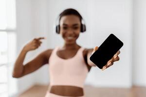 Sports App Concept. Fit young African lady holding and showing smartphone with sporty empty screen for mock up template, pointing at isolated gadget display, blurred background, selective focus photo