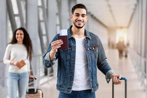 Flight Booking Service. Happy Arab Man Posing With Passport And Tickets At Airport, Handsome Smiling Middle Eastern Guy Standing With Luggage While Waiting For Flight In Terminal, Copy Space photo