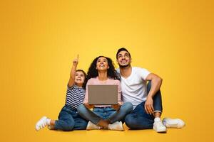 Great Offer. Joyful Arab Family Of Three With Laptop Pointing Up At Copy Space Above Their Heads, Interested Middle Eastern Parents And Little Daughter Looking At Free Place For Advertisement photo