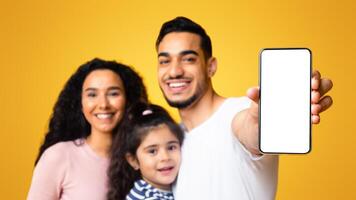 Happy middle eastern family of three showing blank smartphone with white screen for mockup, excited arab parents and little daughter recommending new mobile app, standing on yellow background photo