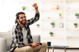 Emotional middle-eastern bearded man playing game with joystick at home, sitting on couch in living room, having fun at weekend, copy space. Home entertainment, leisure, domestic lifestyle photo