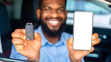 Car Renting App. Happy Black Man Showing Key And Smartphone With Blank White Screen Through Window While Sitting In His Automobile In Dealership Center Or Rental Office, Closeup Shot, Mockup photo