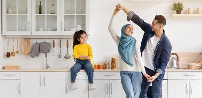 Portrait Of Happy Arab Family Of Three Having Fun In Kitchen Interior, Cute Smiling Little Middle Eastern Girl Sitting On Table And Watching Her Parents Dancing, Enjoying Time At Home Together photo