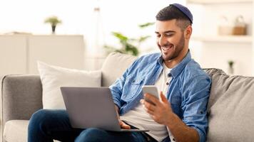 Portrait of smiling jewish man in yarmulke sitting on couch, holding and using mobile phone, reading text message. Cheerful productive guy working on laptop computer, free copy space photo