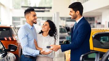 Smiling sales manager giving happy middle-eastern young family key from brand new auto, side view. Emotional arabic couple buying car at luxury delarship salon. Car purchasing concept photo