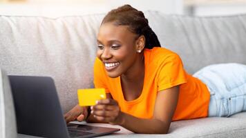 Cheerful Afro woman shopping online with credit card and laptop, lying on sofa at home. Young black lady surfing web, buying goods at internet store, ordering products remotely photo