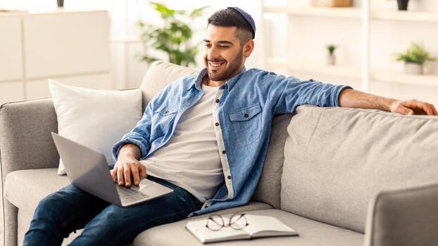 sonriente joven judío hombre relajante en cómodo sofá en vivo habitación utilizando moderno computadora portátil, contento milenario masculino descansando en acogedor sofá a hogar, acecho película o en línea, disfrutando fin de semana foto