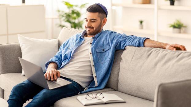 persona de libre dedicación jornada laboral. sonriente milenario hombre empleado estudiante trabajando estudiando desde hogar sentado en sofá en vivo habitación. masculino utilizando ordenador portátil comprometido en electrónico papeleo haciendo distante tarea foto