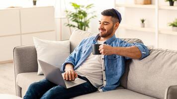 Positive smiling jewish man in yarmulke sitting on the couch, holding coffee mug and working on laptop. Guy checking email. Handsome young male using computer while drinking a cup of hot tea photo