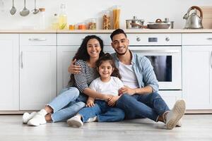 Beautiful middle-eastern family of three sitting on floor at kitchen, hugging and cheerfully smiling at camera, copy space. Happy arab father, mother and daughter spending time together at home photo
